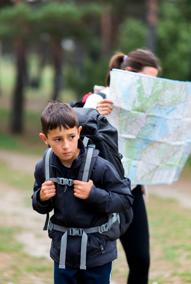 Imagine simbolică: un copil cu ghiozdan greu, în spatele unui părinte care ține harta drumului – reprezentând iubirea transformată în control și povara succesului proiectat asupra copilului.